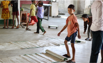 A Santal boy in an orange shirt playing on rolled mats with other children indoors