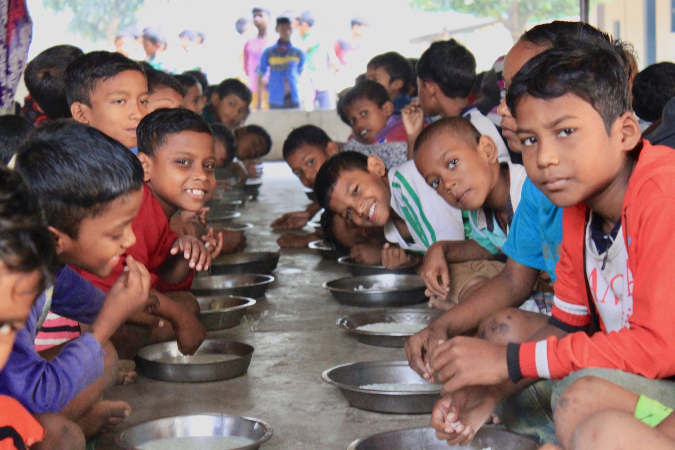 Santal children seated in rows with metal plates for a meal in the dining hall